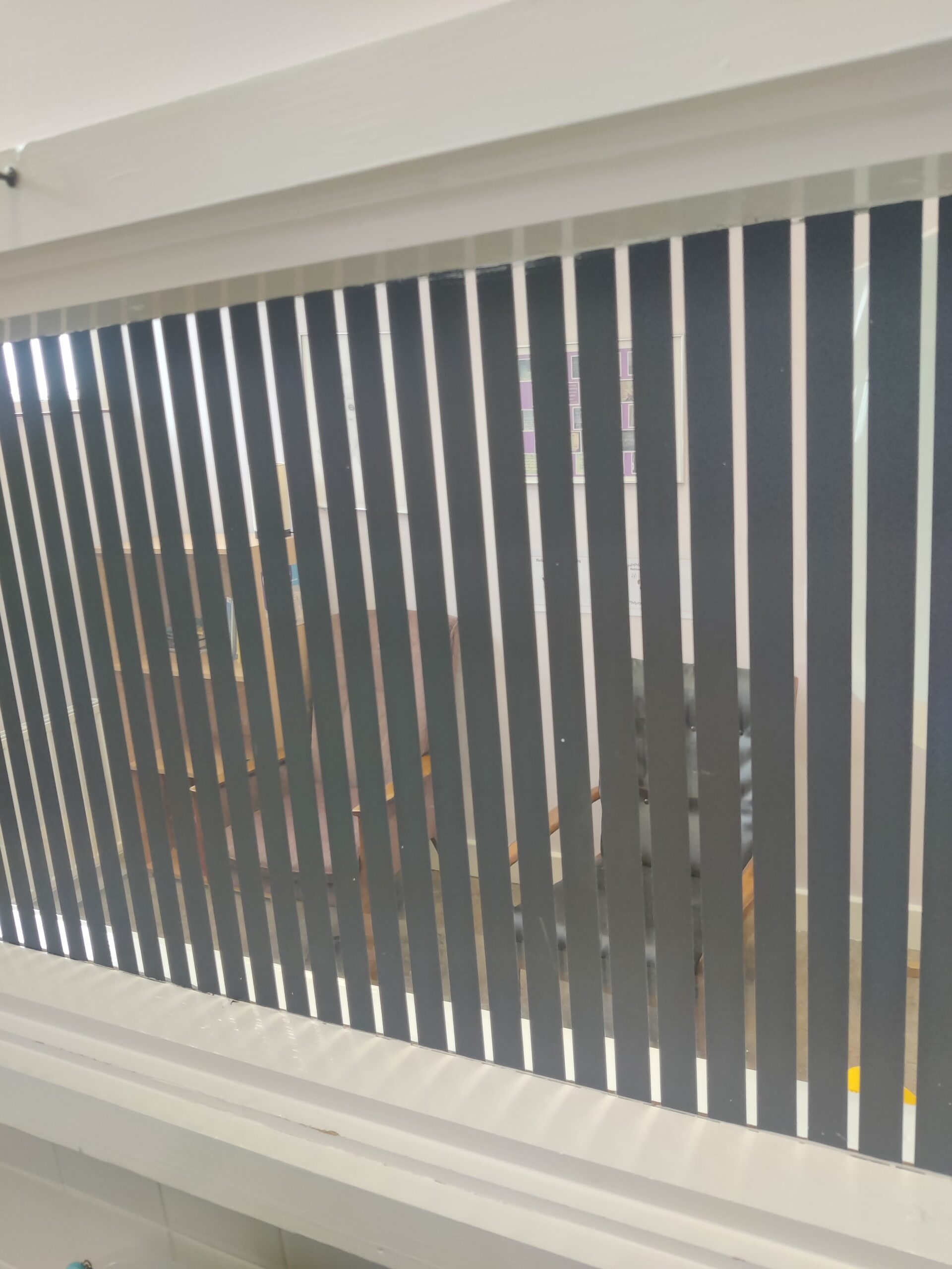A window with vertical slats. The room behind the window has chairs and bookshelf in it.