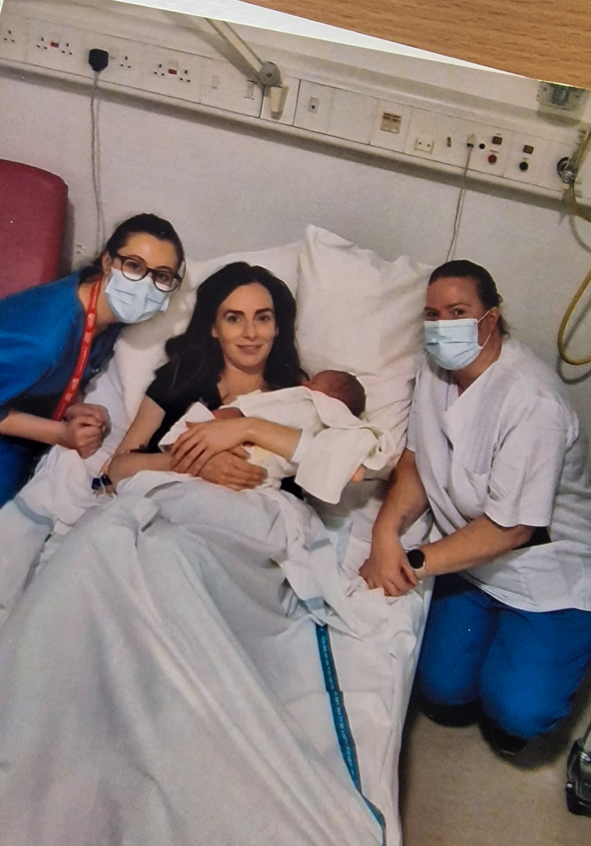 Two midwives crouching either side of a women holding a newborn baby, laying in a hospital bed.