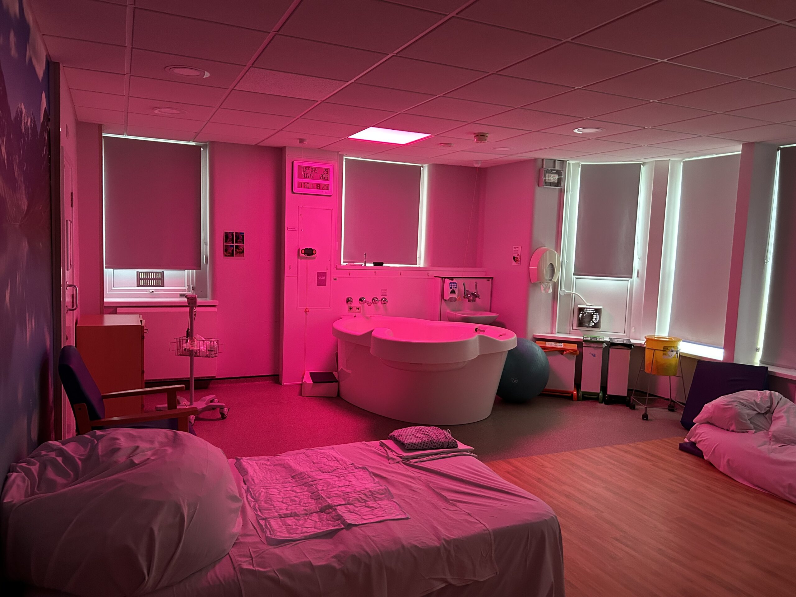 A birth centre room with a bed, a bedside table, and a bathtub bathed in pink light.